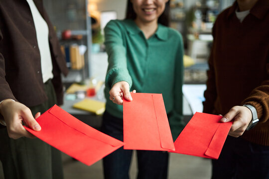Young Asian businessmen and businesswomen holding red envelopes and standing together in office, smiling and extending hands forward, partial bodies visible, focus on gesture