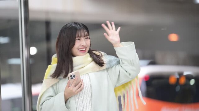 冬の年末年始の帰省、駅のホームやバスターミナルなどでスマホを持つかわいいストールを巻いた女性、手を振るバイバイのポーズ動画
