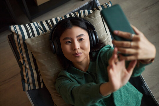 Portrait of young adult Asian woman lying on couch wearing headphones holding smartphone taking selfie smiling slightly relaxed expression modern office environment