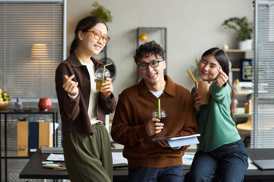 Group of young Asian men and women standing together in modern office, smiling and holding drinks, making hand gestures while interacting and looking at camera, casual business setting