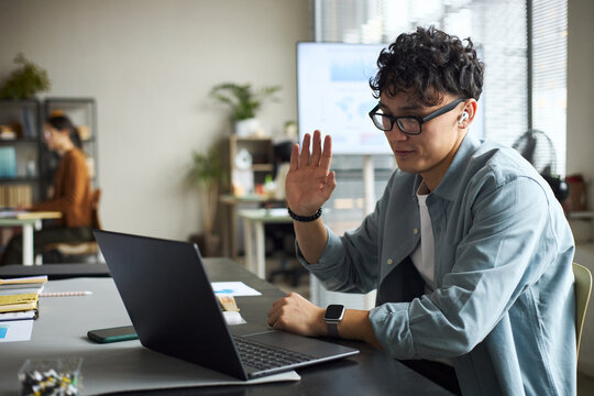 Young Asian man wearing glasses sitting at desk using laptop, raising hand while participating in video conference call in modern office, businesswoman working in background