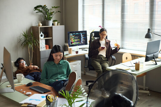 Young Asian businessmen and businesswomen working in modern office, young Asian woman sitting at desk using computer, young Asian man relaxing with headphones, young Asian woman using smartphone