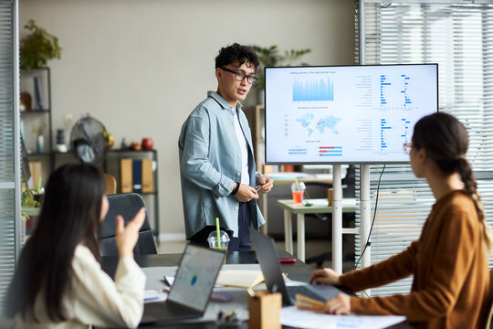 Young Asian man presenting data on digital screen while young Asian businesswoman and young Asian businessman, sitting at table working on laptops in modern office meeting