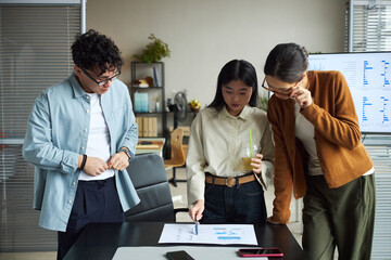 Young Asian man and two young Asian women standing around office table analyzing printed charts and graphs, discussing business strategy, one woman holding drink, digital devices visible