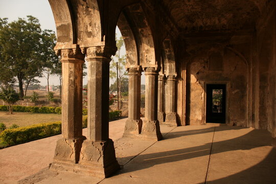 A colonnade of weathered, historic stone pillars forming an arched gallery (veranda) in an ancient Indian structure, lit by warm sunlight. Heritage architecture.