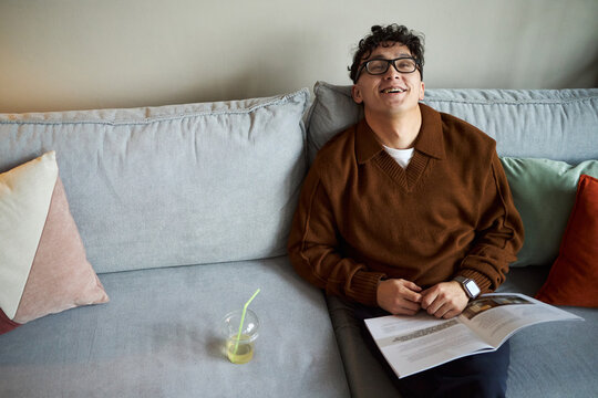 Young adult Asian man sitting on sofa smiling and holding open magazine, wearing glasses and smartwatch, relaxing during break in modern office, plastic cup with straw on cushion nearby