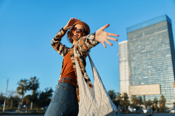 Woman with smile in city, candid lifestyle moment wearing sunglasses and tote during golden hour glow; authenticity and mindful living captured in emotional storytelling urban portrait.