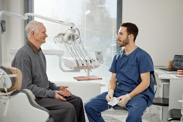 Senior Caucasian man sitting in dental chair, talking with young adult Caucasian male dentist wearing scrubs and gloves in modern dental office with dental equipment visible