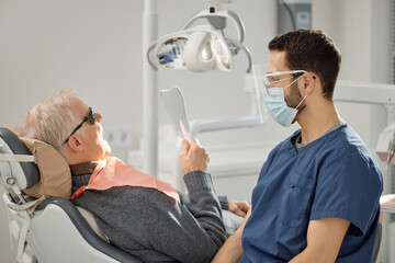 Senior Caucasian man reclining in dental chair holding mirror while young adult male dentist wearing mask and protective glasses sitting nearby during dental consultation