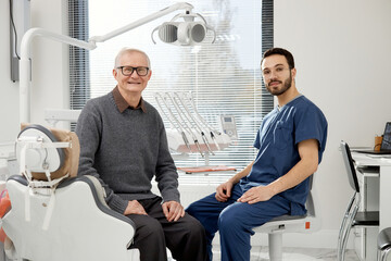 Senior Caucasian man sitting in dental chair, smiling at camera with young adult male dentist in scrubs sitting beside him in modern dental office during appointment