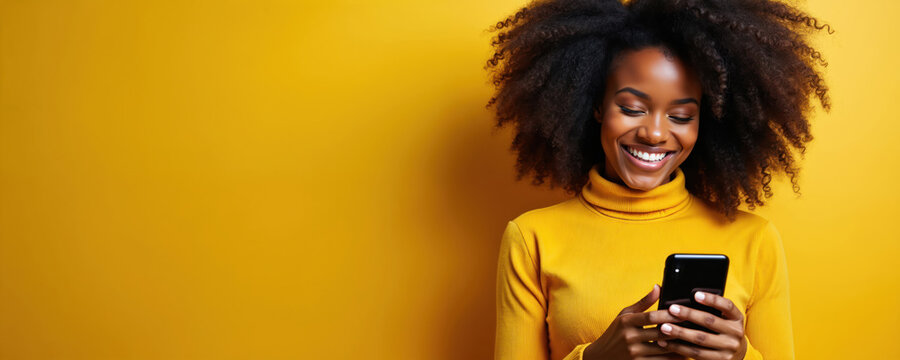Happy young african american woman with curly hair holds smartphone, smiles. Wears yellow turtleneck against plain yellow backdrop. Empty copy space for ad text. Joyful lady using mobile phone in