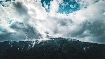 Storm clouds loom above a dark evergreen mountain ridge as rising mist threads through the dense forest. Patches of blue sky break the gloom. A dramatic wilderness scene with moody weather today.
