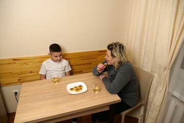 Family moment with mother and son sitting at table, drinking tea, eating cookies, and enjoying warm conversation together indoors.