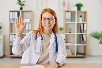 Friendly smiling female doctor looking at camera waving hand having online video call or conference...