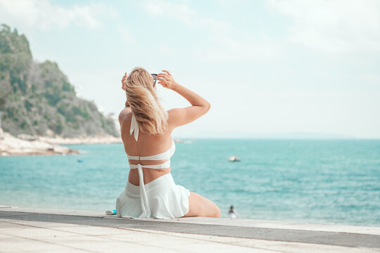 A young woman in a white bikini sits on the sea wall, hand in hair, gazing at the turquoise ocean with lush cliffs behind, capturing pure summer beach bliss. Tropical paradise, vacation vibe.