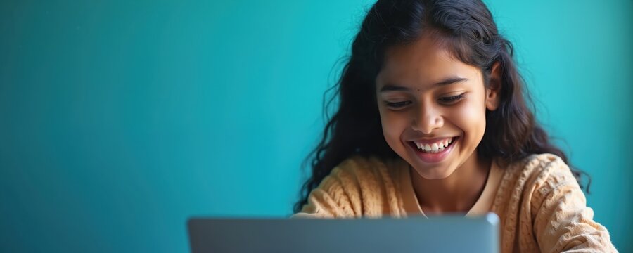 Young Indian girl smiles while using tablet computer. She studies online learning remotely. Bright blue background suggests digital education. Focus on happy student, technology access, future skills.