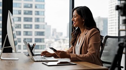 Happy young businesswoman smiling and multitasking with smartphone & laptop in a modern office. Professional, productive female entrepreneur working in a contemporary high-rise workspace with city vie - Powered by Adobe