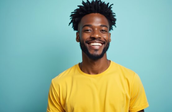 Young Black man with afro hair wears yellow shirt smiles brightly at camera. Studio portrait against plain pastel blue background conveys positive emotion and confidence for lifestyle concepts.