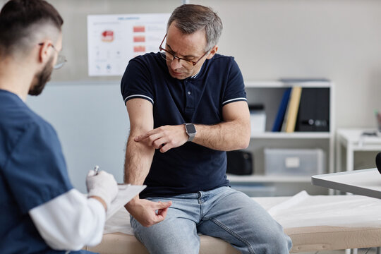 Middle aged Caucasian man sitting in dermatologist office, showing irritated skin on forearm to young adult male doctor wearing medical gloves holding clipboard during consultation