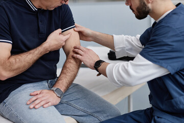 Middle aged Caucasian man sitting while male healthcare professional examining upper arm during dermatology appointment, both focusing on skin condition in clinical setting