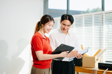 Two Asian coworkers working together on project planning, reviewing documents and using digital devices in modern office.