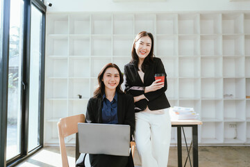 Two businesswomen work together at a laptop, smiling as they collaborate in an office with stacks of documents on the desk.