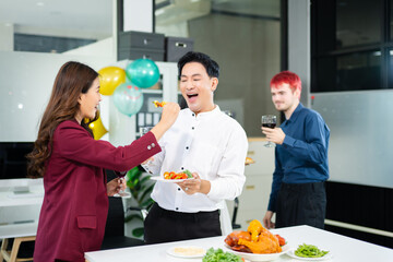 Cheerful coworkers share laughs as one playfully feeds a skewer during a festive office lunch with wine, food, and laughter, celebrating together in a modern workplace.
