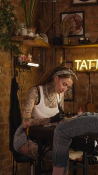 Vertical side view shot of professional young Caucasian female tattoo artist tattooing on leg of her unrecognizable client in cozy modern studio with warm dim lighting
