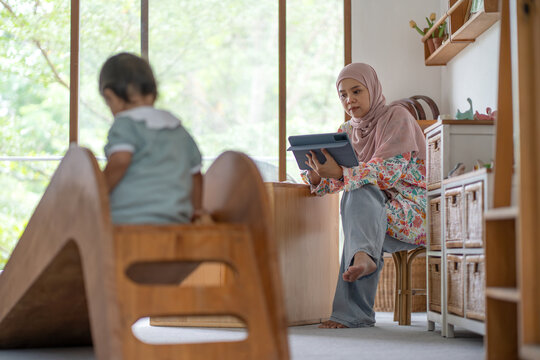 Indonesian Muslim Woman Remote Working on Tablet with Daughter Playing Indoors