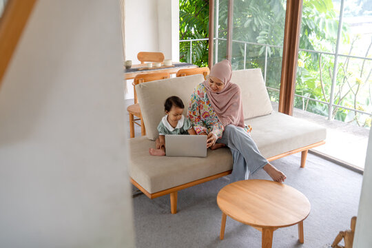 Indonesian Muslim Woman Remote Working at Home with Daughter on Sofa