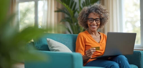 Elderly woman happily uses credit card for online shopping on laptop. Mature lady makes internet purchase on sofa, smiles at screen. Technology for seniors. Paying bills easily at home.
