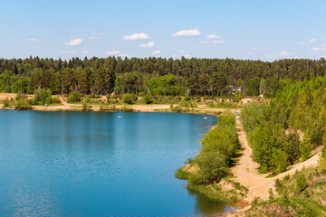 Vibrant blue quarry lake contrasts with lush green forest and sandy shoreline under a bright summer sky, perfect for outdoor recreation and nature lovers