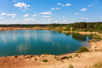 Obraz premium Stunning turquoise water in a sandy quarry lake surrounded by lush green forest under a bright blue sky with fluffy clouds