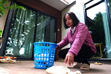 A woman is squatting next to a blue laundry basket, picking up clothes that have fallen onto the...