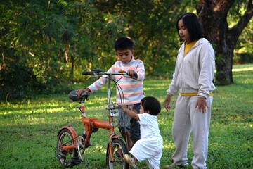 Fototapeta premium A woman is squatting next to a bicycle, helping a small child sit securely in the front basket, while an older child is seated on the saddle.