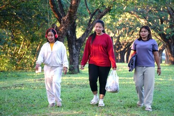 Three women are walking across a grassy park area surrounded by trees, with the woman in the middle carrying a plastic bag with items.