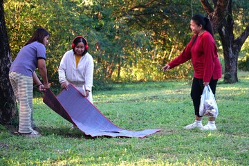 Two women are cooperating to unroll a picnic mat on the grass, while their friend stands nearby...