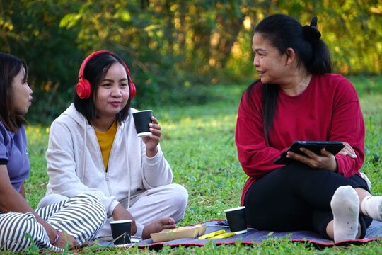 Three women sitting on the grass for a picnic, the woman in the middle is holding a paper cup of beverage, while the woman in the red shirt is holding a tablet.