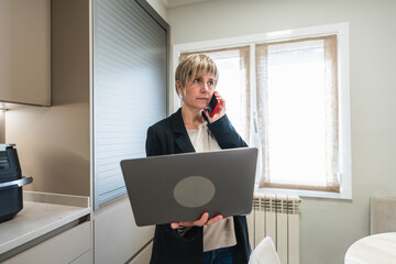 Woman managing a phone call and laptop, balancing work responsibilities in her home kitchen during remote work