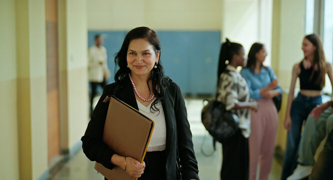 Indian mature tutor gen x adult woman hold file folder walk indoor college hall space pupil boy girl do talk spend break time happy smiling female lady carry bag go inside office corridor work place