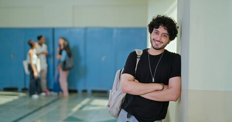 Naklejka premium Indian happy young adult gen z proud man male look camera cross arm standing campus area. Cool pupil boy curly hair wear bag fold hand staring cam do casual pose enjoy new good day time indoor place