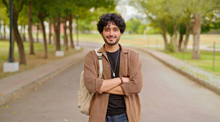 Indian happy young adult gen z man male look camera cross arm standing outdoor area. Smart cool proud pupil boy guy wear bag fold hand staring cam do casual pose enjoy new day outside place