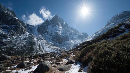 Snowy alpine mountains under a bright sun, a rocky trail in the foreground and low shrubs along the hillside. Concept Snowy alpine peaks, Bright sunlit slopes, Rocky trail foreground