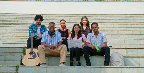 Indian young adult gen z guys male man lady female group team sitting on stair look camera spend break time. Happy teen pupil boy girl staring cam do casual pose enjoy new day have fun joy place