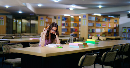 Indian young adult gen z teen age student wear casual glasses sitting on desk table do self study read novel indoor library focused girl look book hold pencil prepare exam make skill task notes © Raushan_films