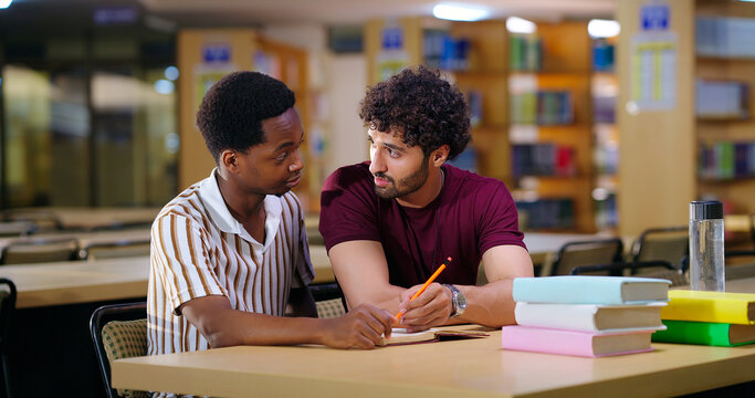 Indian two teen male sitting on desk table do study talk new project task work share plan ideas young adult gen z boys man guys pair hold pencil look book read exam notes task indoor library place