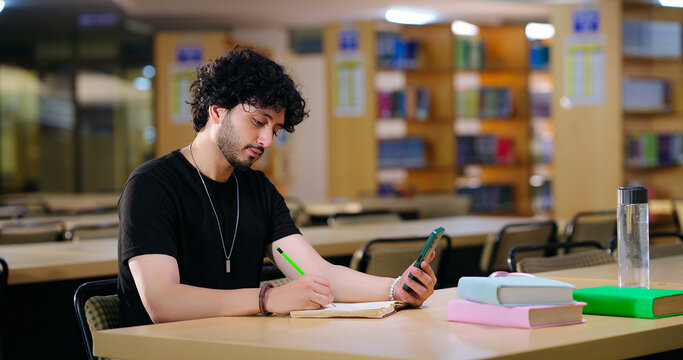 Indian young adult gen z man pupil sitting on desk table use mobile hold pencil take exam notes book smart boy male guy look phone screen do self study in good WIFI e learning new task work at library