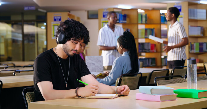 Indian Asian young adult gen z male man calm peace mind student sitting desk table listening music hold pencil write exam notes smart boy do self study read book prepare topics indoor library busy day