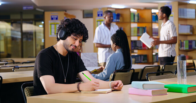 Indian Asian young adult gen z male man calm peace mind student sitting desk table listening music hold pencil write exam notes smart boy do self study read book prepare topics indoor library busy day