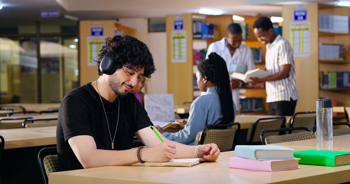 Indian Asian young adult gen z male man calm peace mind student sitting desk table listening music hold pencil write exam notes smart boy do self study read book prepare topics indoor library busy day